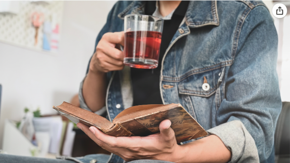 Person sitting on a couch holding a book and a glass of red restoraid.