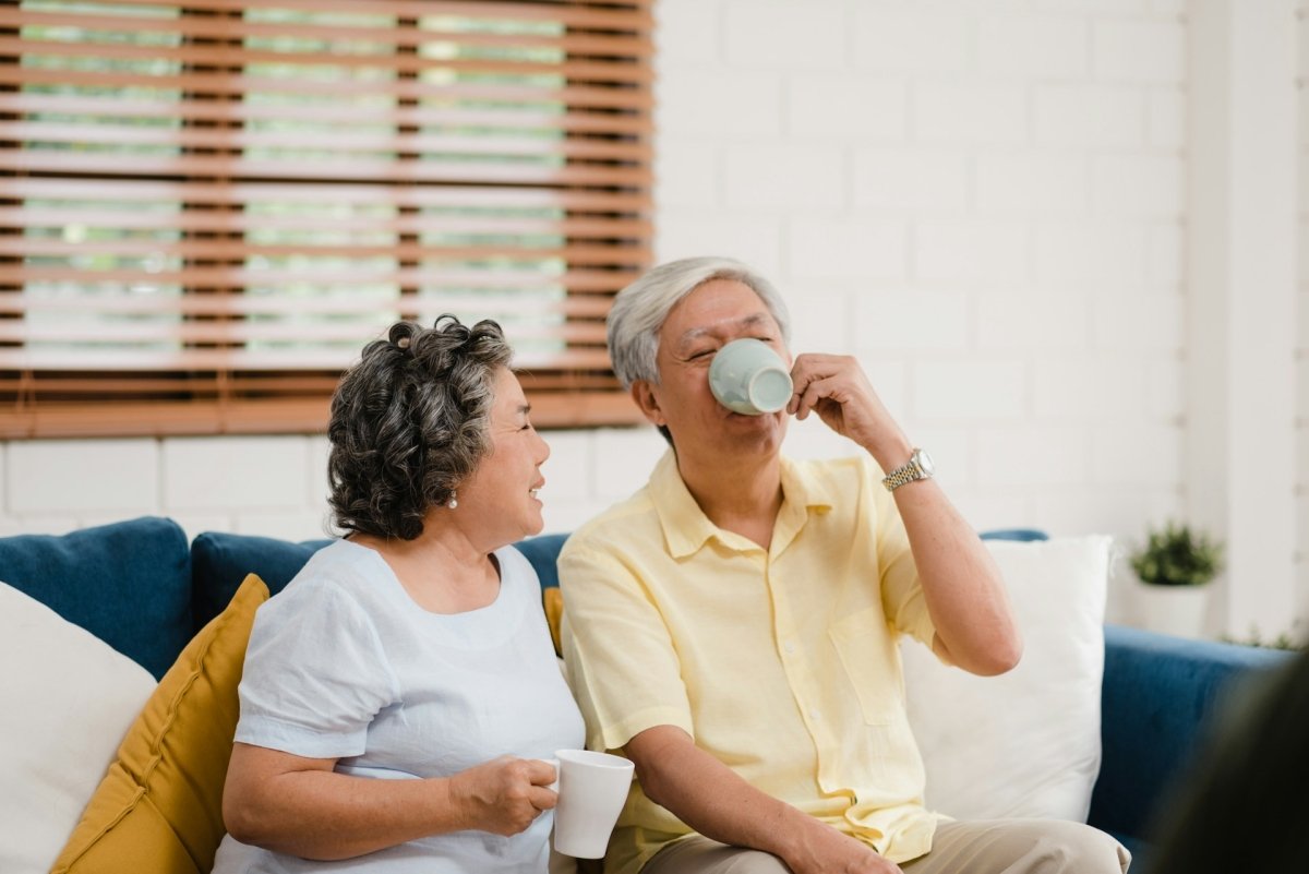 Two elderly people sitting on a couch, one holding a cup and the other sipping from a mug.