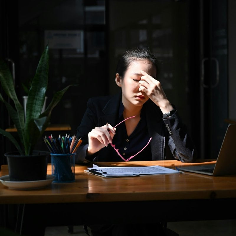 Person sitting at a desk with a laptop, holding their head in a dimly lit room.