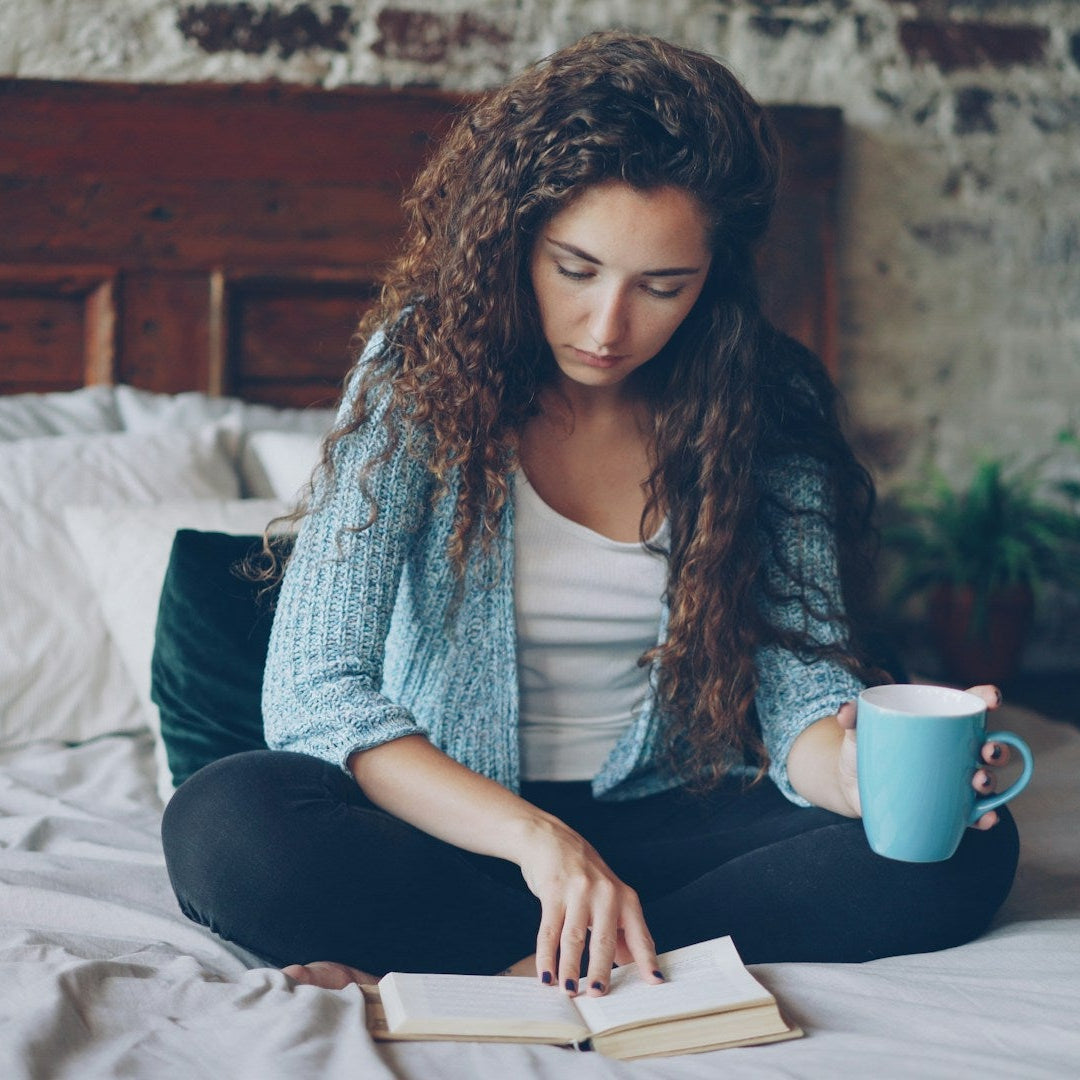 Woman reading a book on a bed with a cup in her hand