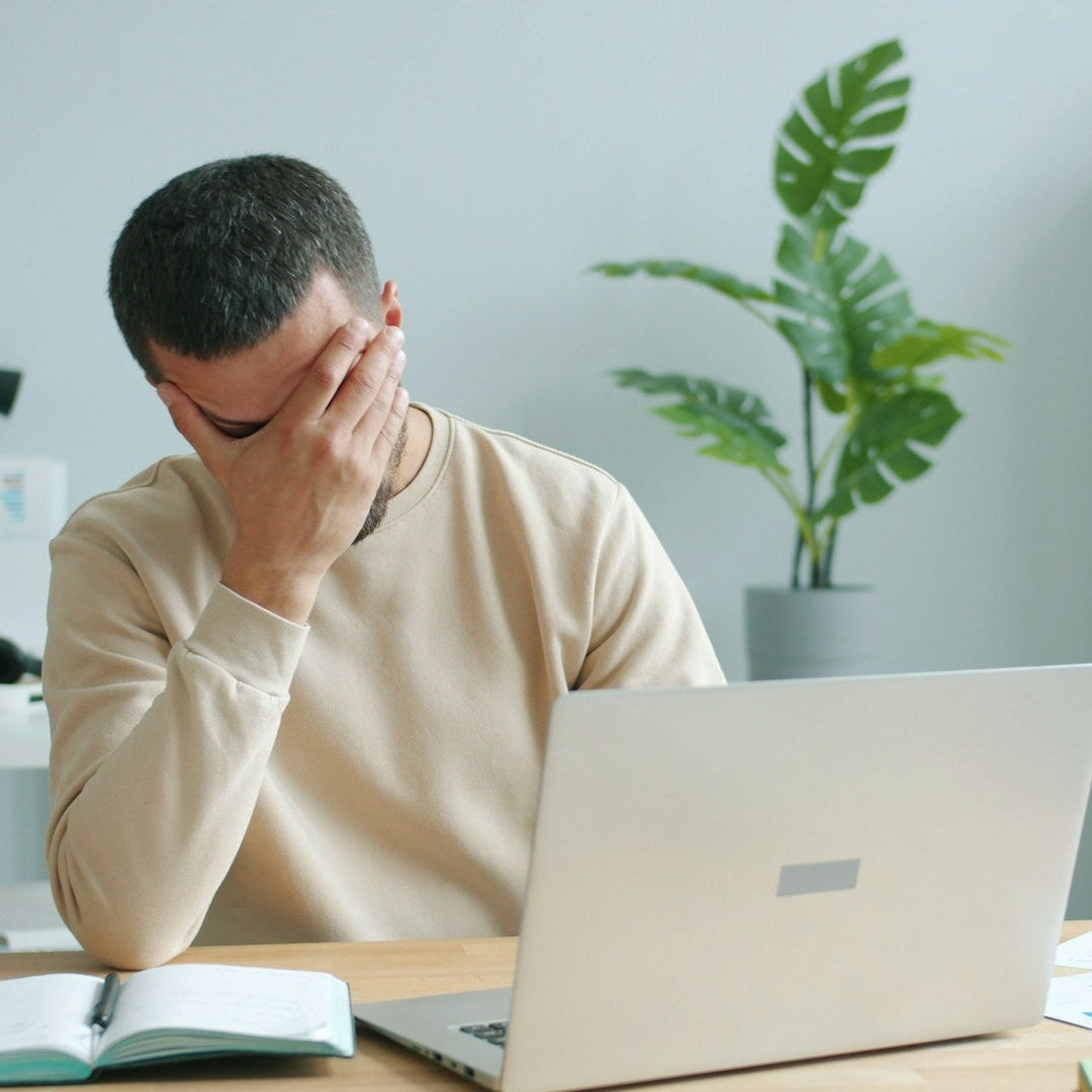 Person sitting at a desk with a laptop, holding their head in a room with a plant and shelves.